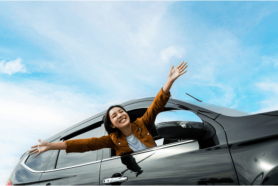 Femme souriante sortant de la fenêtre d’une voiture noire, bras levés sous un ciel bleu
